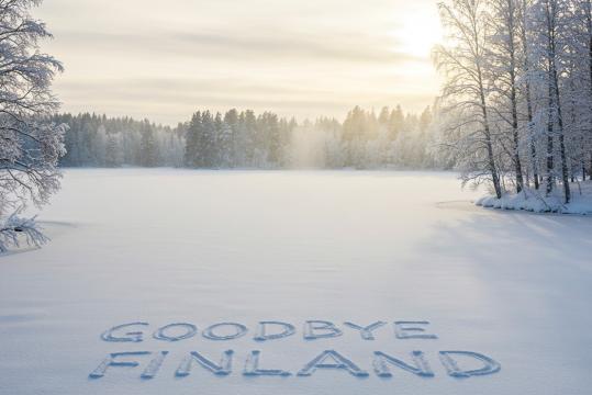 A winter landscape featuring a frozen lake surrounded by snow-covered trees. The sun is low in the sky, casting a soft golden glow through the mist. In the foreground, "GOODBYE FINLAND" is carefully imprinted in the fresh snow, creating a striking contrast against the untouched white surface.